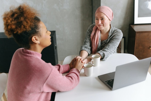 Two women sitting at a table and holding hands next to two coffee mugs and a laptop.